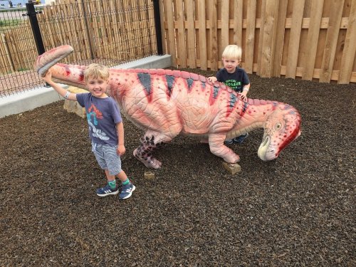 Two children play beside a large dinosaur sculpture in a playground, smiling and posing for the camera.