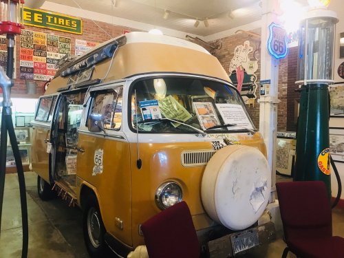 A vintage yellow camper van displayed indoors, surrounded by retro signs and a vintage gas pump.