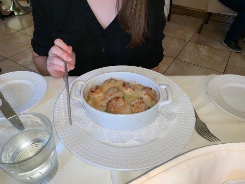 A person holds a fork next to a dish of baked scalloped potatoes on a white plate at a restaurant table.