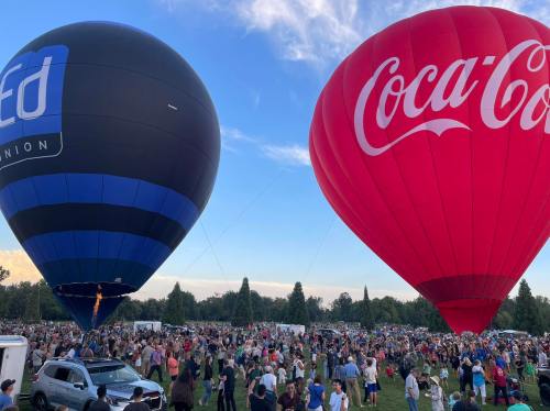 Two large hot air balloons, one black with "Credit Union" and the other red with "Coca-Cola," above a crowd in a park.