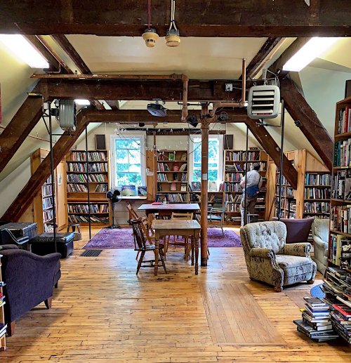 Cozy library interior with wooden beams, bookshelves, a table, and seating areas. A person is organizing books.