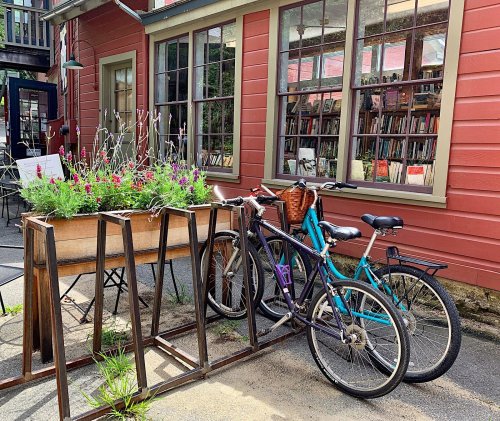 Bicycles parked next to a flower planter outside a quaint building with large windows and a book collection inside.