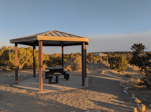 A wooden gazebo with a picnic table, surrounded by desert vegetation and a clear sky.