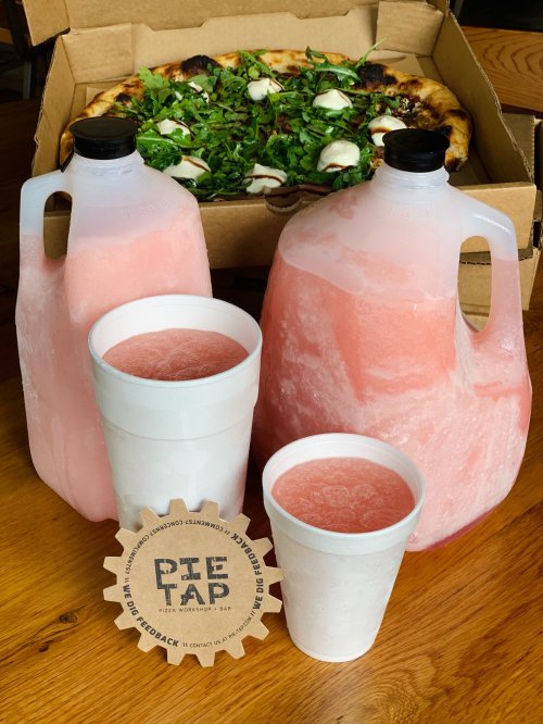 Two large jugs of pink drink, two cups, and a pizza box with a pizza topped with greens and cheese in the background.