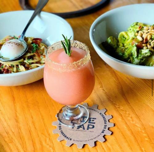 A pink cocktail garnished with rosemary sits on a wooden table, surrounded by bowls of pasta and salad.
