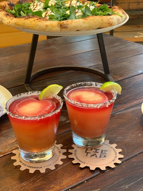 Two colorful cocktails with lime slices on a wooden table, alongside a pizza in the background.
