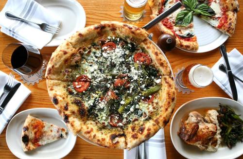 A table spread with various pizzas, plates of food, and drinks, featuring a large pizza topped with greens and pepperoni.