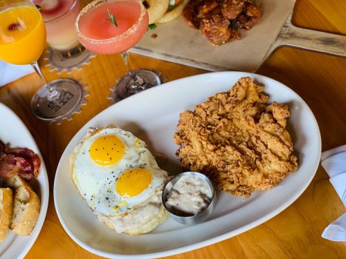 A plate with two fried eggs, crispy fried chicken, and a small cup of dipping sauce, with drinks in the background.