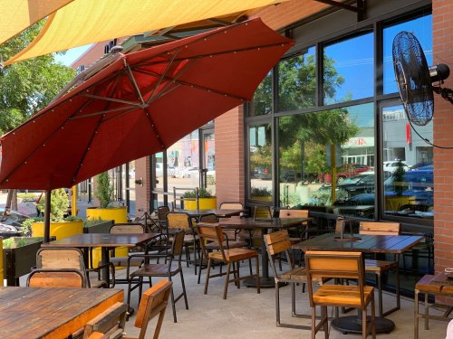Outdoor café seating with wooden tables and chairs, shaded by a large red umbrella, beside large windows.