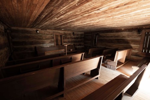 Interior of a rustic wooden building with empty pews and a simple altar, illuminated by natural light.