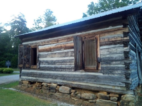 A rustic log cabin with wooden windows, surrounded by trees and gravel path.