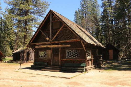 A rustic wooden cabin with a steep roof, surrounded by tall trees and a clear blue sky.