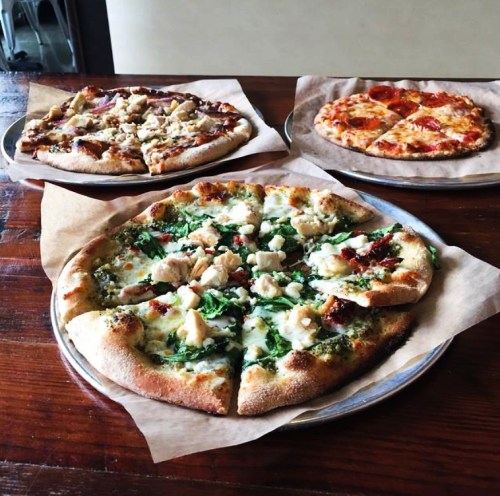 Three pizzas on metal trays: one with chicken and greens, one with pepperoni, and one with a mix of toppings.