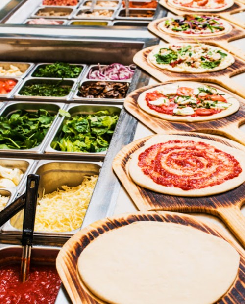 A variety of pizza ingredients and prepared pizzas on wooden boards, with fresh toppings and sauces displayed.