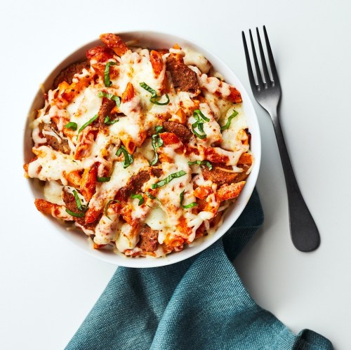 A bowl of baked pasta topped with melted cheese and fresh basil, placed on a teal napkin with a fork beside it.