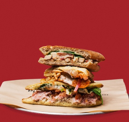 A stacked sandwich with layers of chicken, greens, and toppings on a wooden plate against a red background.