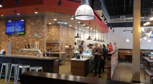 A modern restaurant interior with a brick wall, bar area, and customers waiting near the counter.