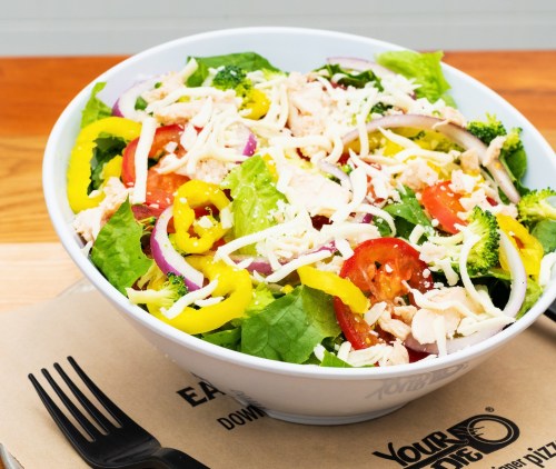 A colorful salad with lettuce, tomatoes, broccoli, peppers, onions, and shredded cheese in a white bowl on a wooden table.