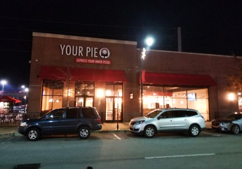 Exterior of a "Your Pie" pizza restaurant at night, featuring large windows and red awnings, with parked cars in front.
