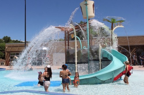 Children and adults enjoy a water slide and splash pad on a sunny day, with water cascading from a large bucket.