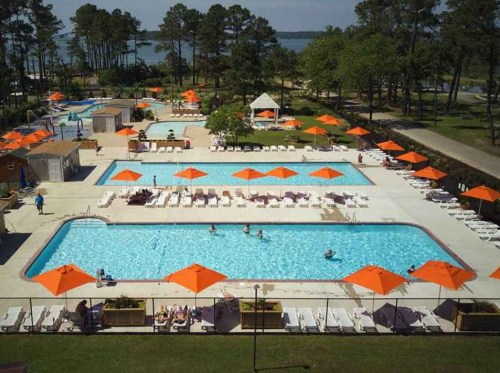 Aerial view of a resort pool area with multiple pools, orange umbrellas, and people enjoying the sunny day.