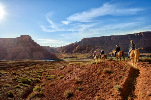 Four riders on horseback traverse a scenic, rugged landscape under a bright blue sky.