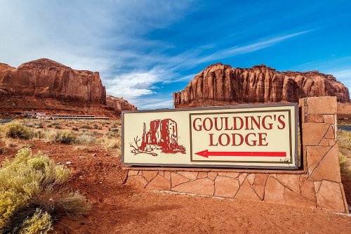 Sign for Goulding's Lodge with red rock formations and blue sky in the background. Arrow points to the left.