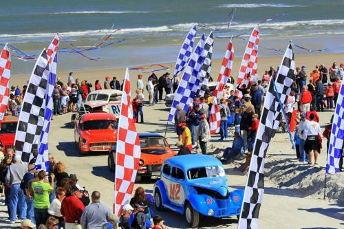 A lively beach event with classic cars, checkered flags, and a crowd of spectators enjoying the festivities.