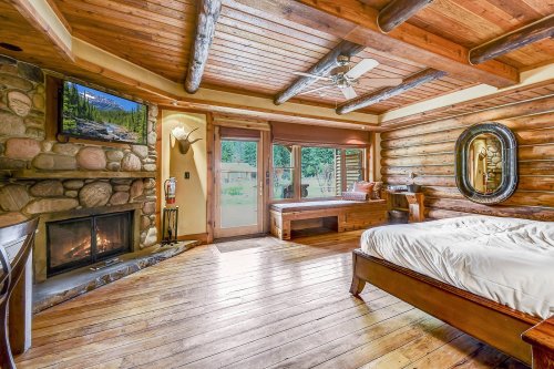 Cozy log cabin bedroom with wooden beams, a stone fireplace, and large windows overlooking nature.