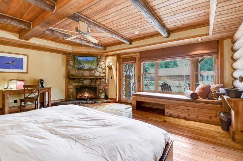 Cozy bedroom with wooden beams, a stone fireplace, and large windows overlooking a serene outdoor view.