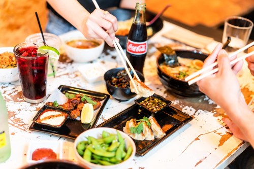 A table filled with various Asian dishes, including ramen, dumplings, and drinks, with hands using chopsticks.