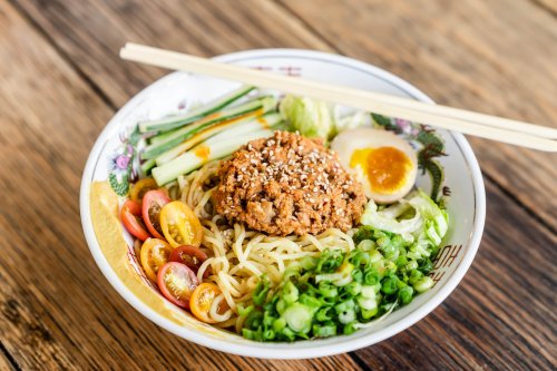A bowl of noodles topped with minced meat, vegetables, cherry tomatoes, and a soft-boiled egg, served with chopsticks.