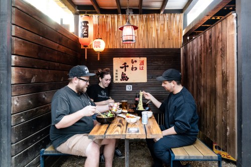 Three people enjoying a meal at a wooden table in a cozy, Japanese-style dining space with lanterns and decor.