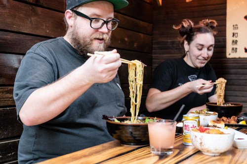 Two people enjoying bowls of noodles at a wooden table, one using chopsticks and the other with a fork.