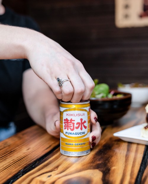 A hand with a ring opens a can of Kikuisui Funaguchi sake on a wooden table with food in the background.