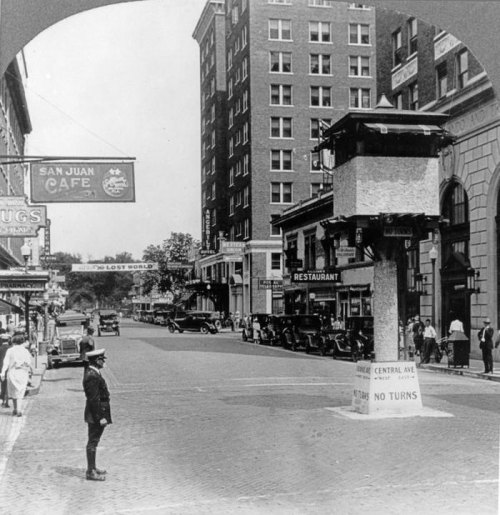 Historic street scene featuring a traffic tower, vintage cars, and pedestrians in a bustling urban setting.
