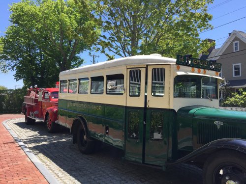 A vintage green bus parked beside a red fire truck on a sunny day, surrounded by trees and a clear blue sky.