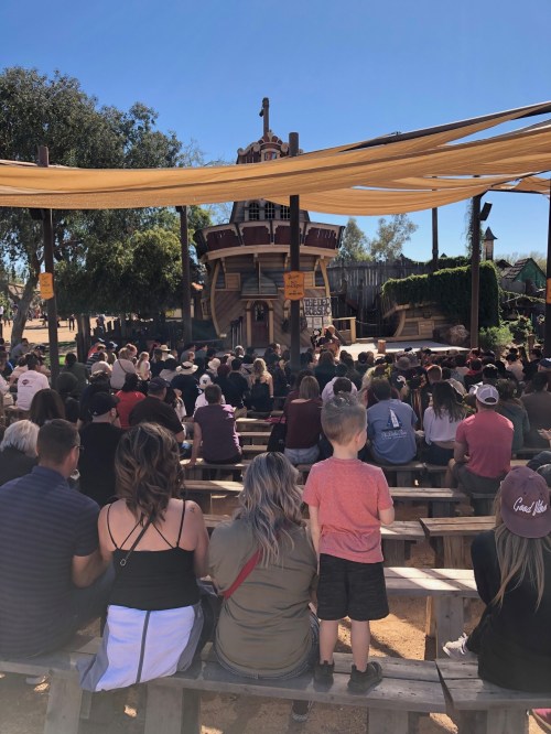 A crowd gathers in an outdoor theater with a wooden stage, shaded by fabric, enjoying a performance on a sunny day.