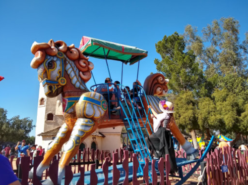 A colorful carousel horse ride with people enjoying it, set against a clear blue sky and trees in the background.