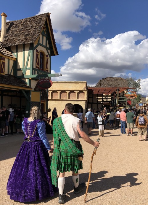 A couple in medieval attire walks through a bustling fair, with shops and people in the background under a blue sky.