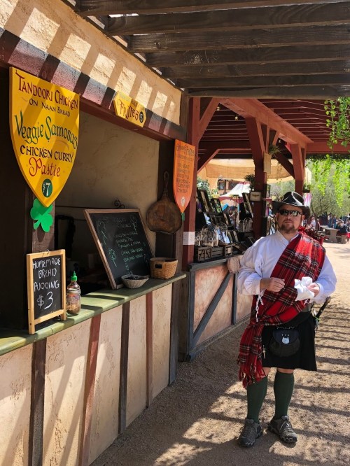 A man in a kilt stands at a food stall with signs for various dishes at a market.