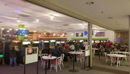 A lively arcade with people playing games, colorful lights, and tables for dining in a spacious setting.