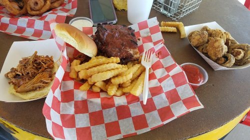 A table with a meal featuring pulled pork, crinkle fries, a sandwich, and fried pickles, all on checkered paper.