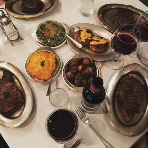 A table set with various dishes including steaks, sides, and red wine, all served on silver platters.