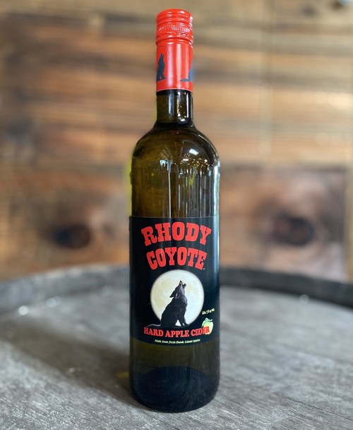 A bottle of Rhody Coyote hard apple cider with a wolf silhouette on the label, set against a wooden background.
