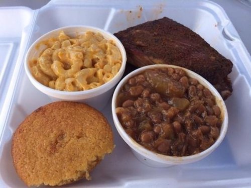 A takeout container with macaroni and cheese, baked beans, cornbread, and a slice of smoked brisket.