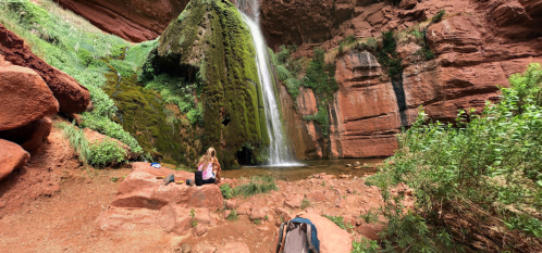 A person sits by a waterfall surrounded by red rock formations and lush greenery in a serene natural setting.