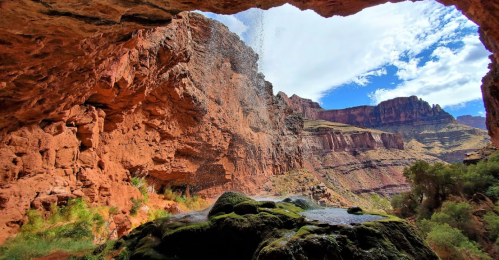 A view from a cave showing a waterfall cascading down red rock formations under a blue sky with scattered clouds.