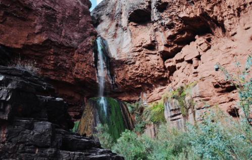 A waterfall cascading down red rock formations, surrounded by lush greenery and rugged terrain.