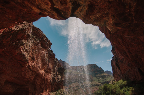 A waterfall cascades through a rocky arch, surrounded by red cliffs and a clear blue sky.